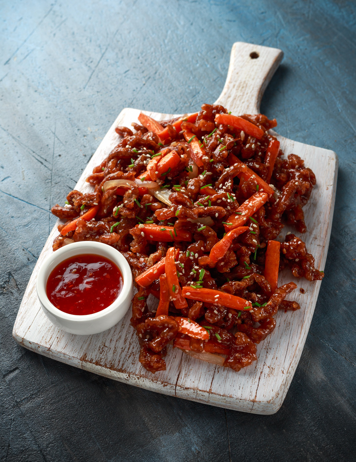Crispy shredded beef with carrots and sweet chilli sauce on white wooden board. Chinese takeaway food